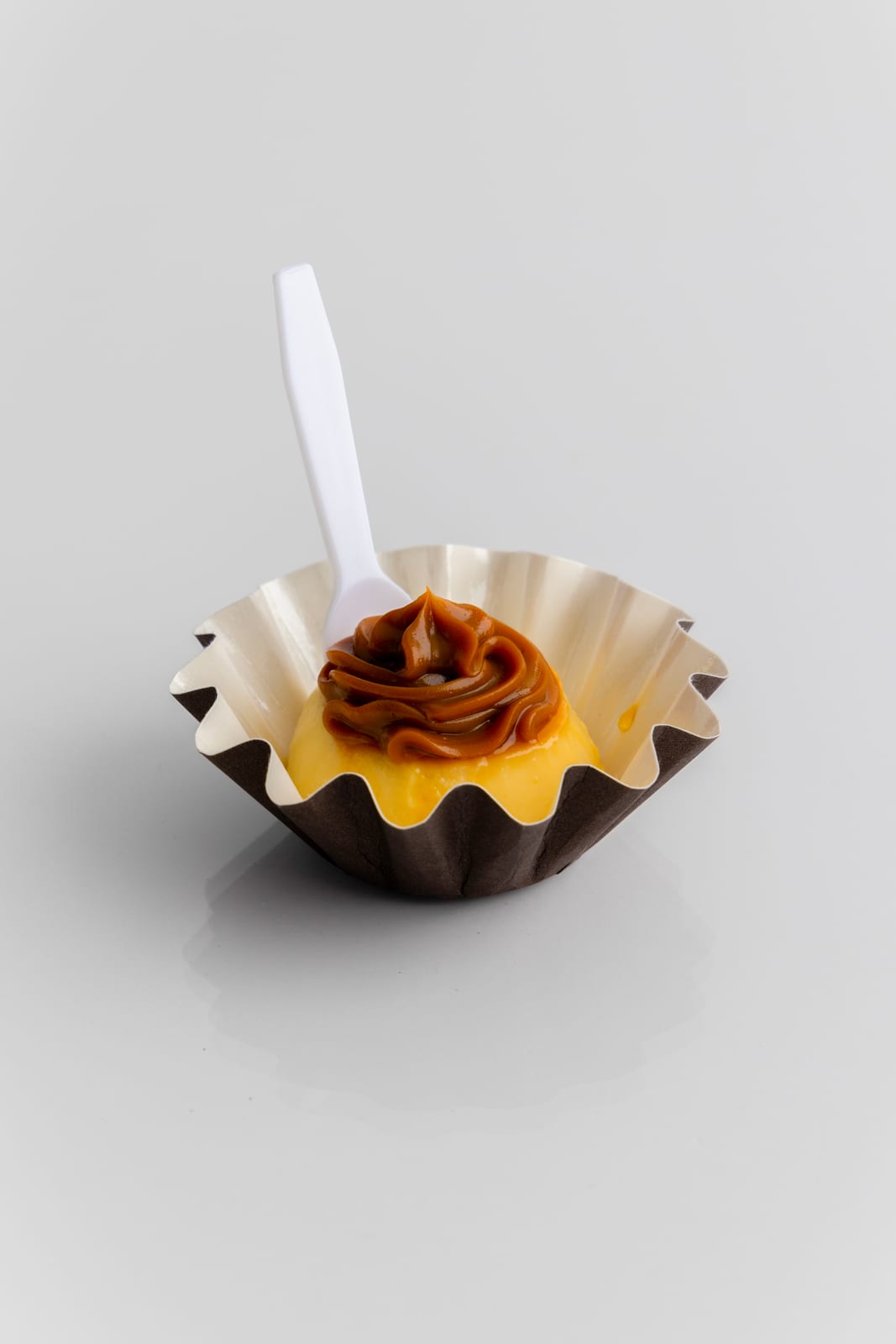 Single mini flan in a fluted paper cup topped with a swirl of dulce de leche, a small white spoon tucked behind it