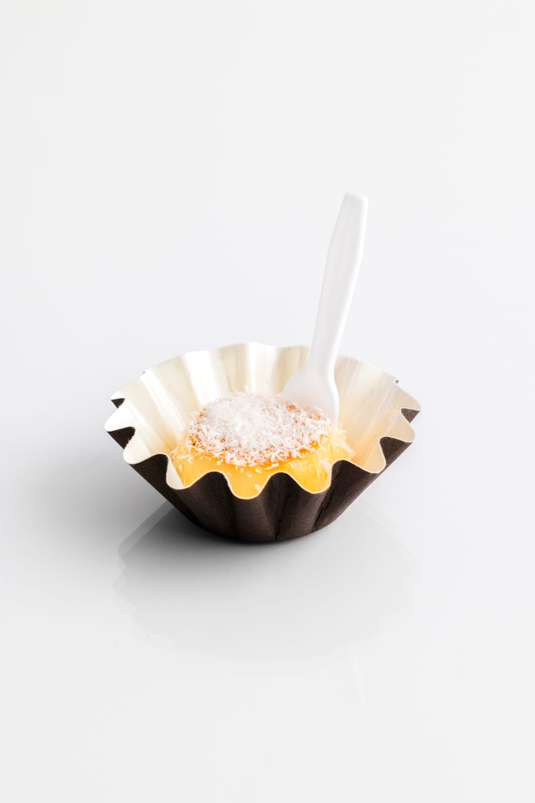 Mini flan in a fluted paper cup covered with shredded coconut, a small white tasting fork standing in it